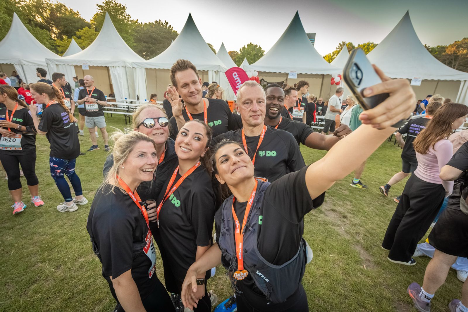 Eine fröhliche Gruppe von Teilnehmenden des B2RUN Dortmund 2025 macht ein gemeinsames Selfie vor weißen Pagodenzelten. Alle tragen schwarze Laufshirts mit dem Logo der Stadt Dortmund („DO“) und orangefarbene Medaillenbänder. Die Stimmung ist ausgelassen, der Teamgeist spürbar – ein Moment voller Stolz und Zusammenhalt nach dem erfolgreichen Lauf.