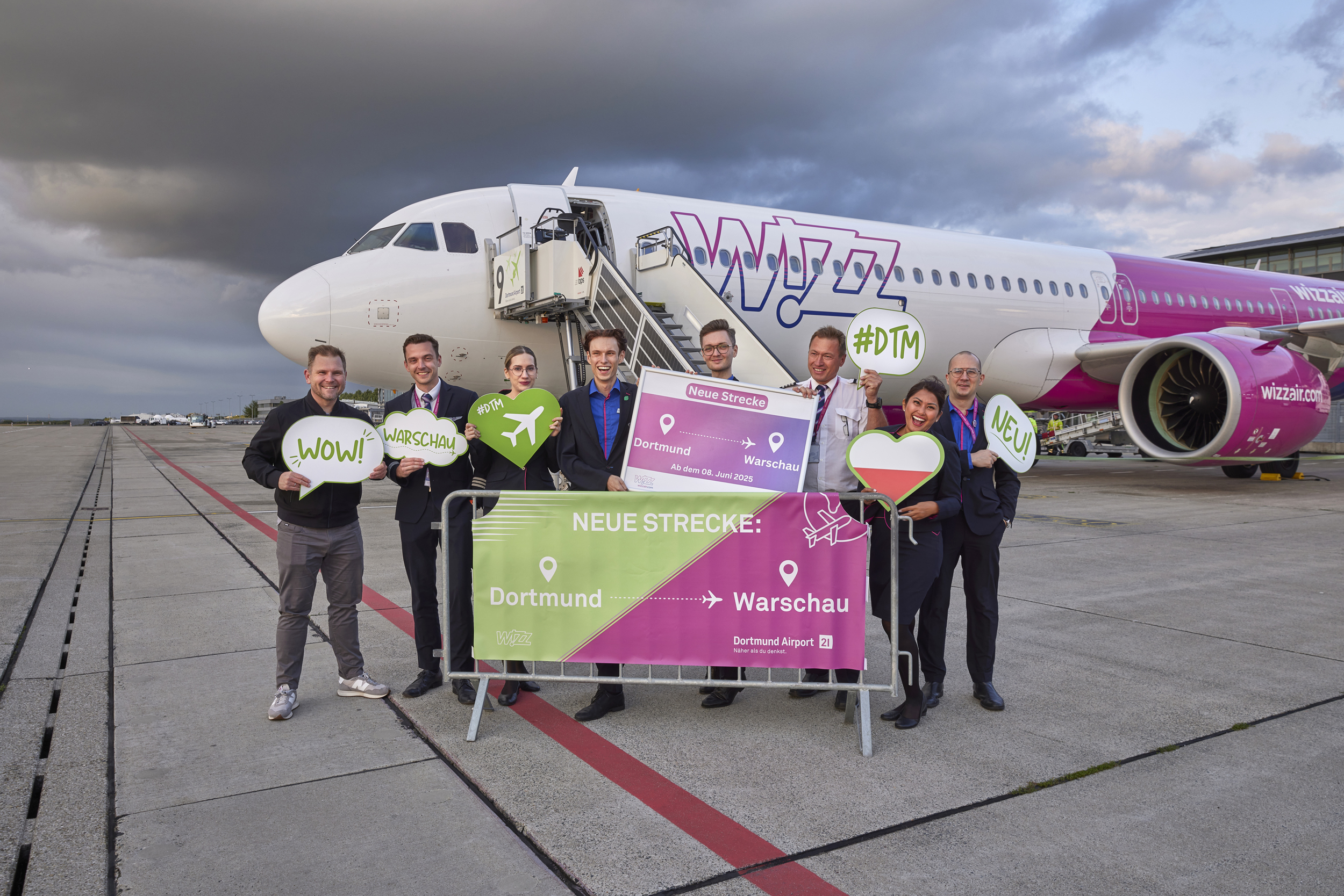 Acht Personen stehen vor einem Wizz Air-Flugzeug auf dem Flugplatz und halten Schilder.