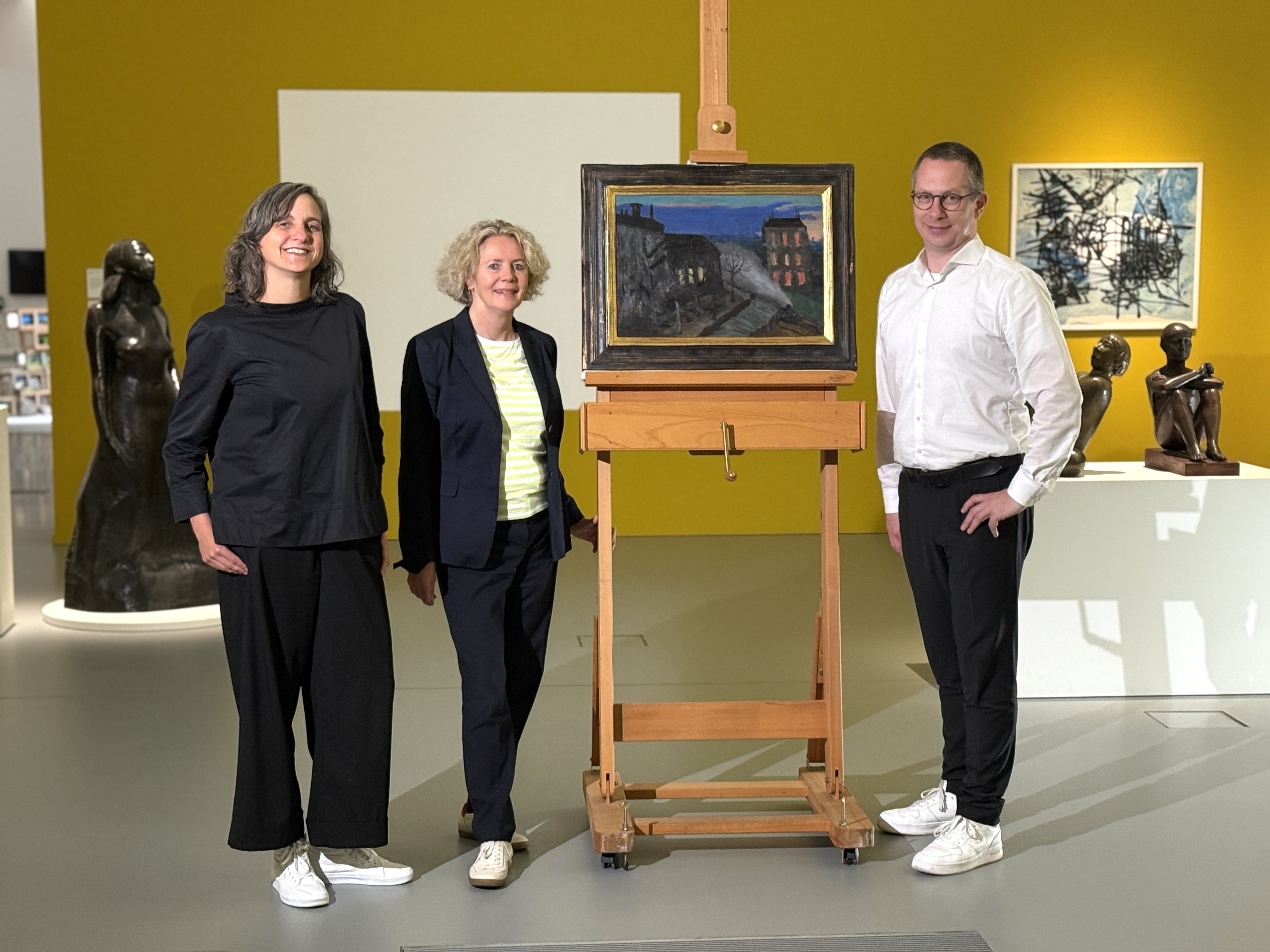 Dr. Nicole Grothe, Direktorin Regina Selter und Benjamin Sieber stehen um das Gemälde, welches zwei Gebäude, einen Weg und Bäume in dunklen Farben zeigt.