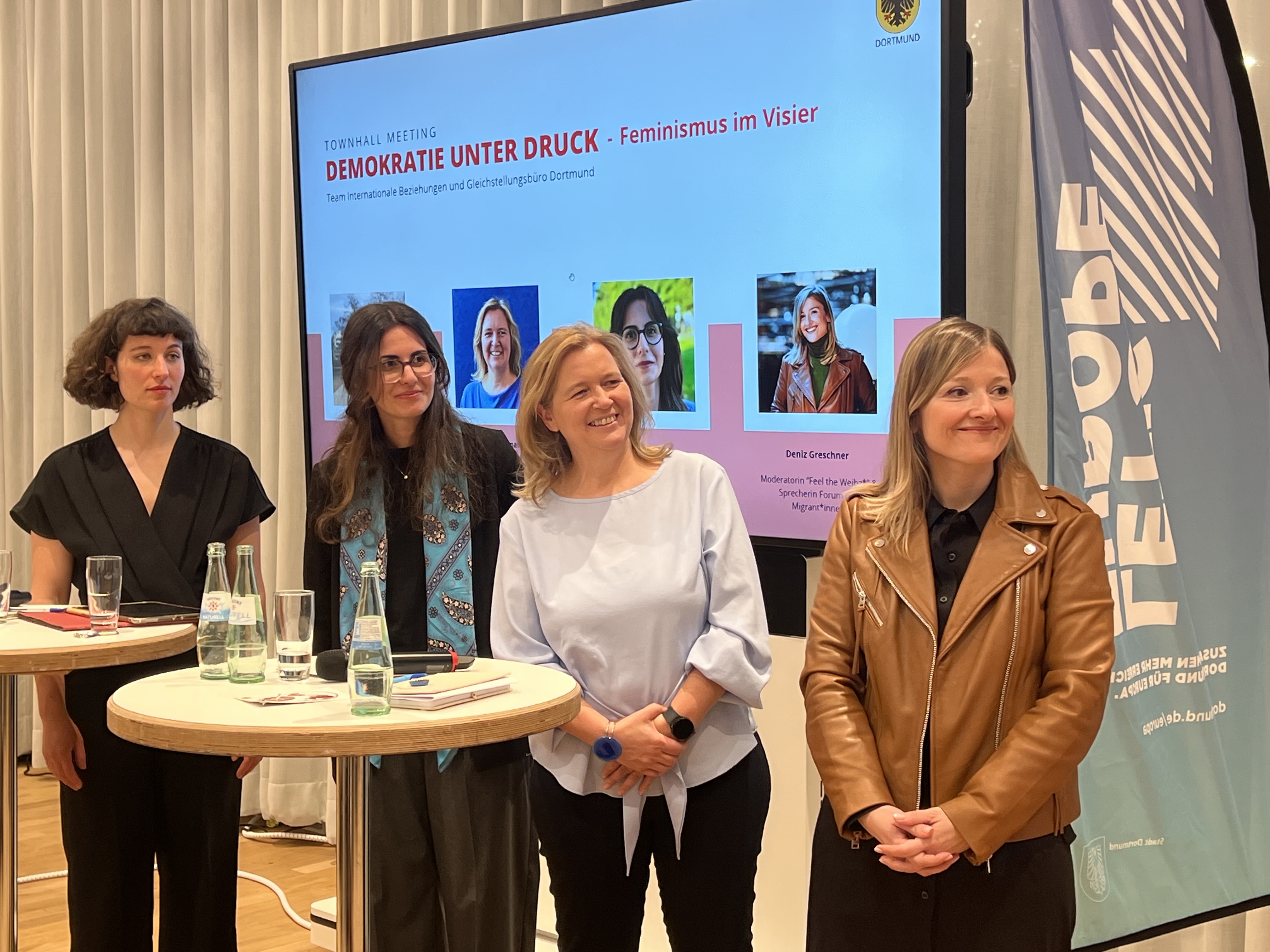 Vier Frauen stehen an einem Stehtisch und lächeln.
